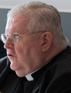 an older man wearing glasses and a priest's robe looks off into the distance