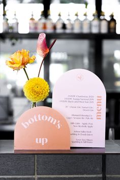 a vase with yellow flowers sitting on top of a counter next to a sign that says bottoms up