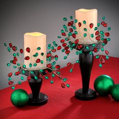 two black vases with red, green and white candles in them on a red tablecloth