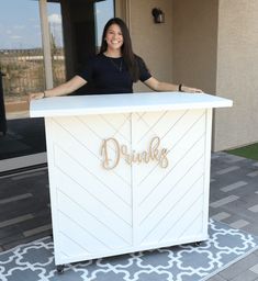 a woman standing behind a white counter in front of a building with the words drinks written on it