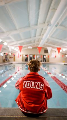 a young boy sitting on the edge of a swimming pool wearing a red jacket that says young