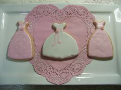 three decorated cookies in the shape of dresses on a white plate with pink doily