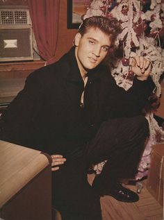 a man sitting next to a christmas tree in front of a red and white flower