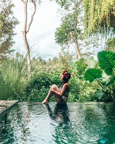 a woman sitting on the edge of a swimming pool surrounded by lush green plants and trees