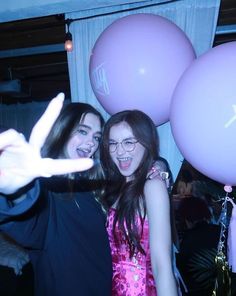 two young women standing next to each other with balloons in front of them and one pointing at the camera