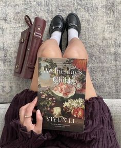 a woman sitting on top of a couch holding a book next to her legs and purse