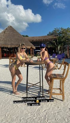 two women in bikinis sitting at a table on the beach with straw huts behind them