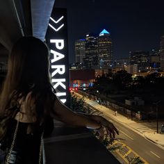 a woman is standing on a balcony looking at the city lights and skyscrapers in the distance