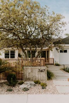 a white house with a wooden fence and trees in the front yard on a sunny day