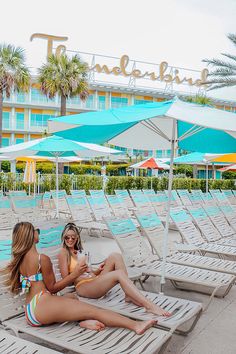 two women sitting on lounge chairs under umbrellas in front of an ocean side hotel