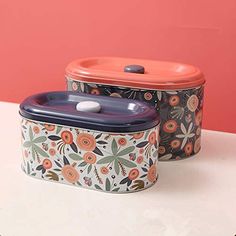 two tins sitting on top of a table with flowers painted on the sides and one has an orange lid