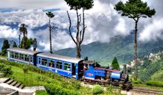 a blue train traveling down tracks next to a lush green hillside covered in clouds and trees