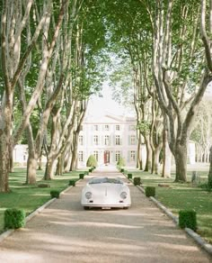 a white car parked in front of a large house surrounded by trees and bushes on either side of the road