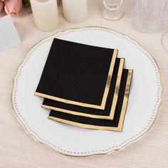 three black and gold napkins on a white plate next to some candles in vases