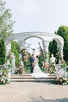 a bride and groom are standing in front of an arch with flowers on the steps