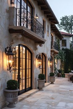 an outdoor patio with potted plants and lights on the side of the house at dusk