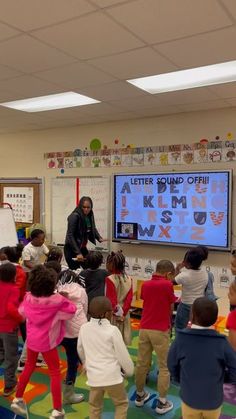 a teacher teaching children in a classroom with a large screen on the wall behind them