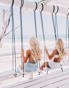 two women sitting on swings looking out at the ocean in front of them and holding onto each other