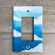 a painted light switch plate on a wooden wall with clouds and blue sky in the background