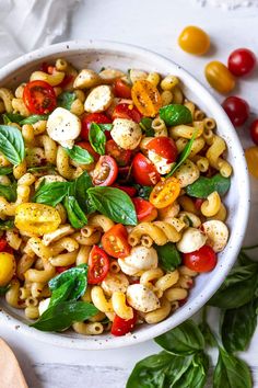 a white bowl filled with pasta salad next to tomatoes, basil and mozzarella