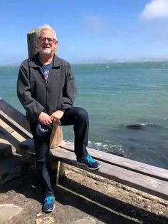 an older man sitting on a wooden bench near the ocean with his feet propped up