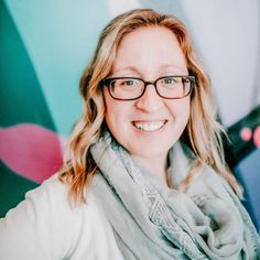 a woman wearing glasses and a scarf smiles at the camera while standing in front of a colorful wall