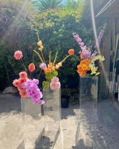 three clear vases filled with colorful flowers on top of a cement floor next to bushes