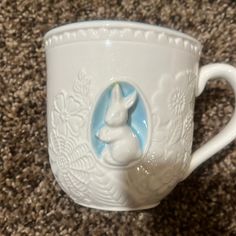 a white coffee cup with a blue bunny in the center on a carpeted floor