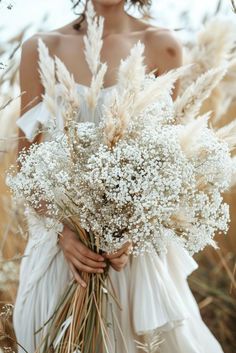 a woman in a white dress holding a bouquet of dried flowers and grass with her hands behind her back