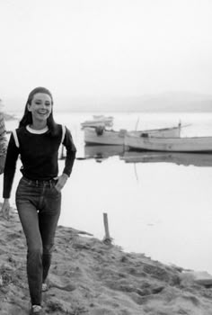 black and white photograph of woman walking on beach with boats in the water behind her