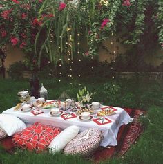 a table with food and drinks on it in the middle of some grass, surrounded by pink flowers