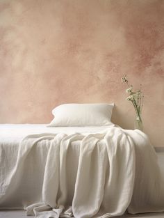 a white bed sitting under a pink wall next to a vase with flowers on it