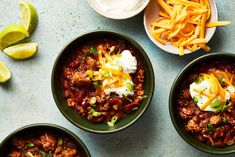 three bowls filled with chili and cheese next to lime wedges on a blue surface