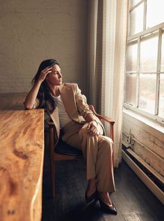 a woman sitting in a chair next to a wooden table with a window behind her