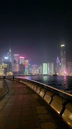 the city skyline is lit up at night from across the water with benches along the edge