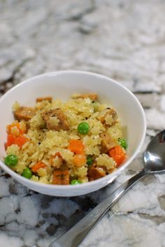 a white bowl filled with rice and veggies next to a spoon on a marble surface