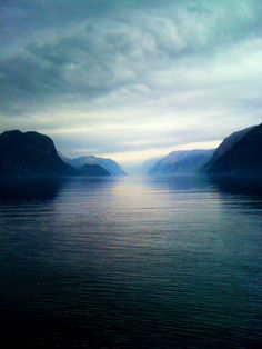 the sun shines brightly through dark clouds over calm water on a cloudy day with mountains in the distance