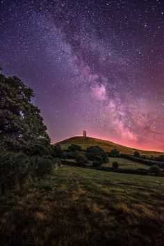 the night sky is filled with stars above a grassy field and tree covered hill in the foreground