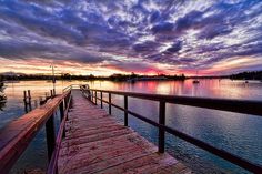 a wooden dock sitting on top of a lake under a colorful sky with clouds above it