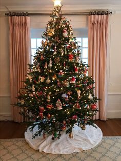 a christmas tree with ornaments on it in front of a window and curtained drapes