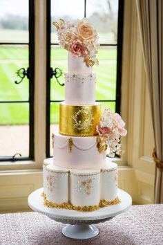 a three tiered wedding cake with gold and pink flowers on the top, in front of a window