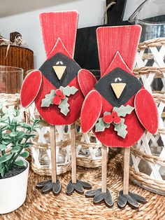 two red wooden birds with holly decorations on them sitting on a wicker table next to a potted plant