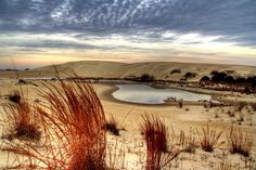 a small lake surrounded by sand dunes and grass in the foreground, under a cloudy sky