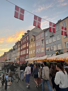 many people are walking on the sidewalk near some buildings and flags flying in the air