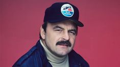 a man with a mustache wearing a blue jacket and hat, against a red background