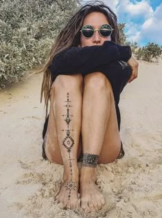 a woman sitting in the sand with her legs crossed and tattoos on her leg,