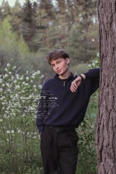 a young man leaning against a tree in the woods with his hand on his hip