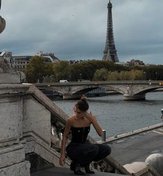 a woman sitting on the edge of a bridge looking at the eiffel tower