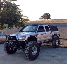 a silver truck parked next to two cars on top of a dirt field with trees in the background