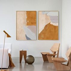 two abstract paintings hang on the wall next to a chair and table in a white room